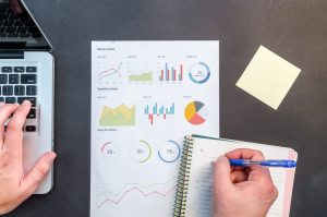 Various graphs on a piece of paper as someone takes notes on a notepad beside a computer